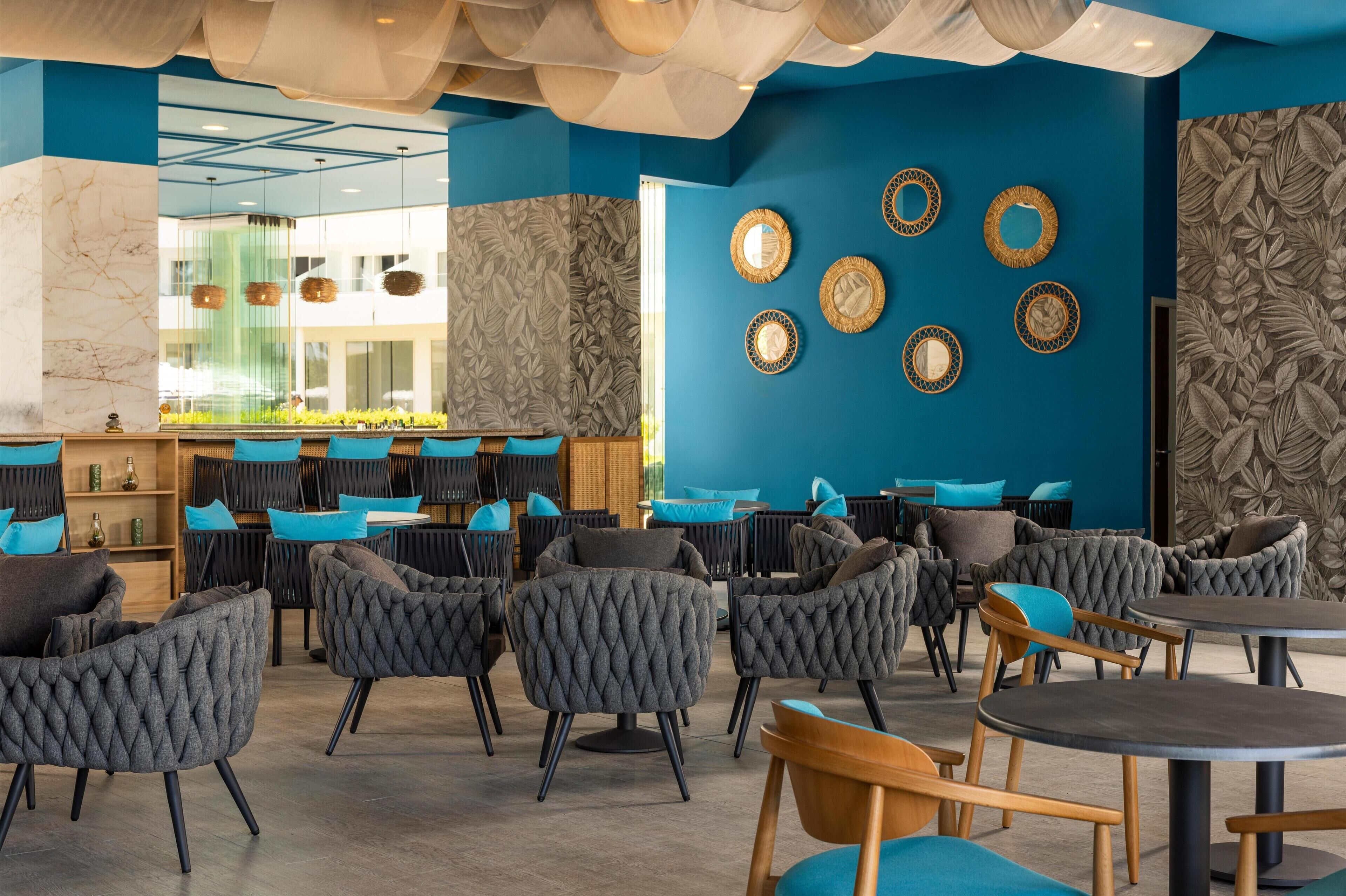 Una cafeteria moderna amb una decoració blau i blanca, amb taules rodones i cadires amb coixins, una zona de bar i decoració mural.