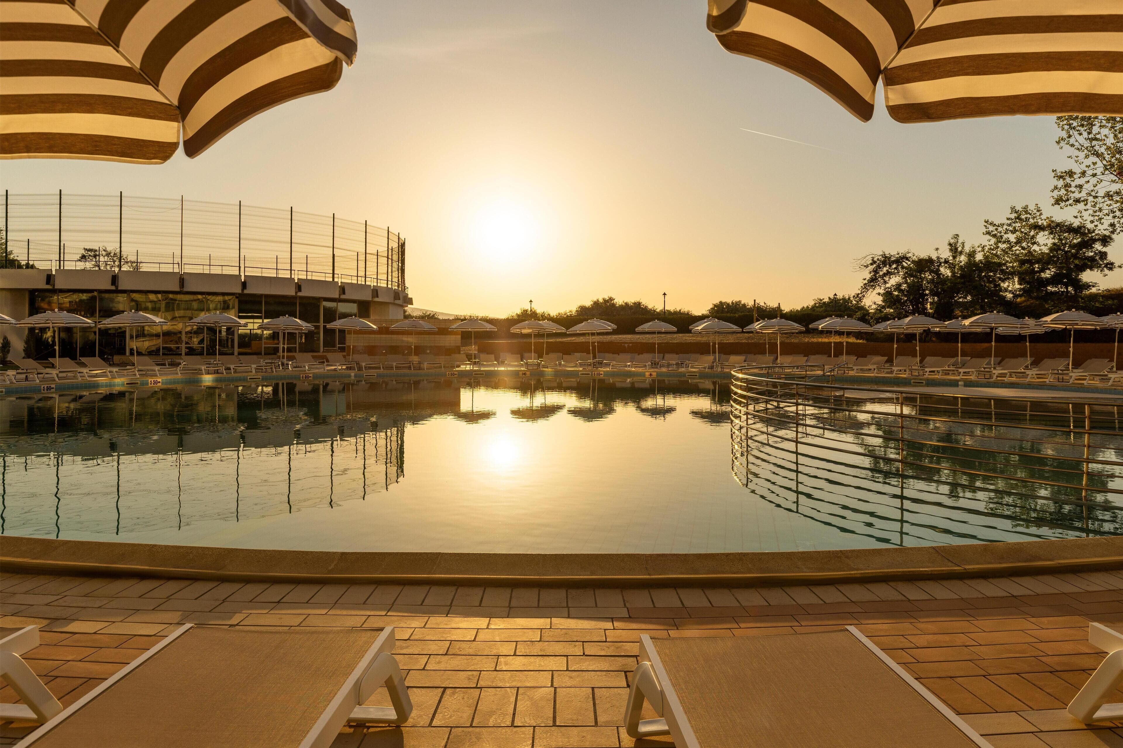 A pool area with lounge chairs and umbrellas, surrounded by a building with a clear view of the sunset.