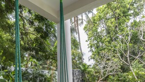 A hammock swing with a white cushion, a stone-tiled floor, and a view of a swimming pool and lush greenery. - Bilin Tree House (Galle)