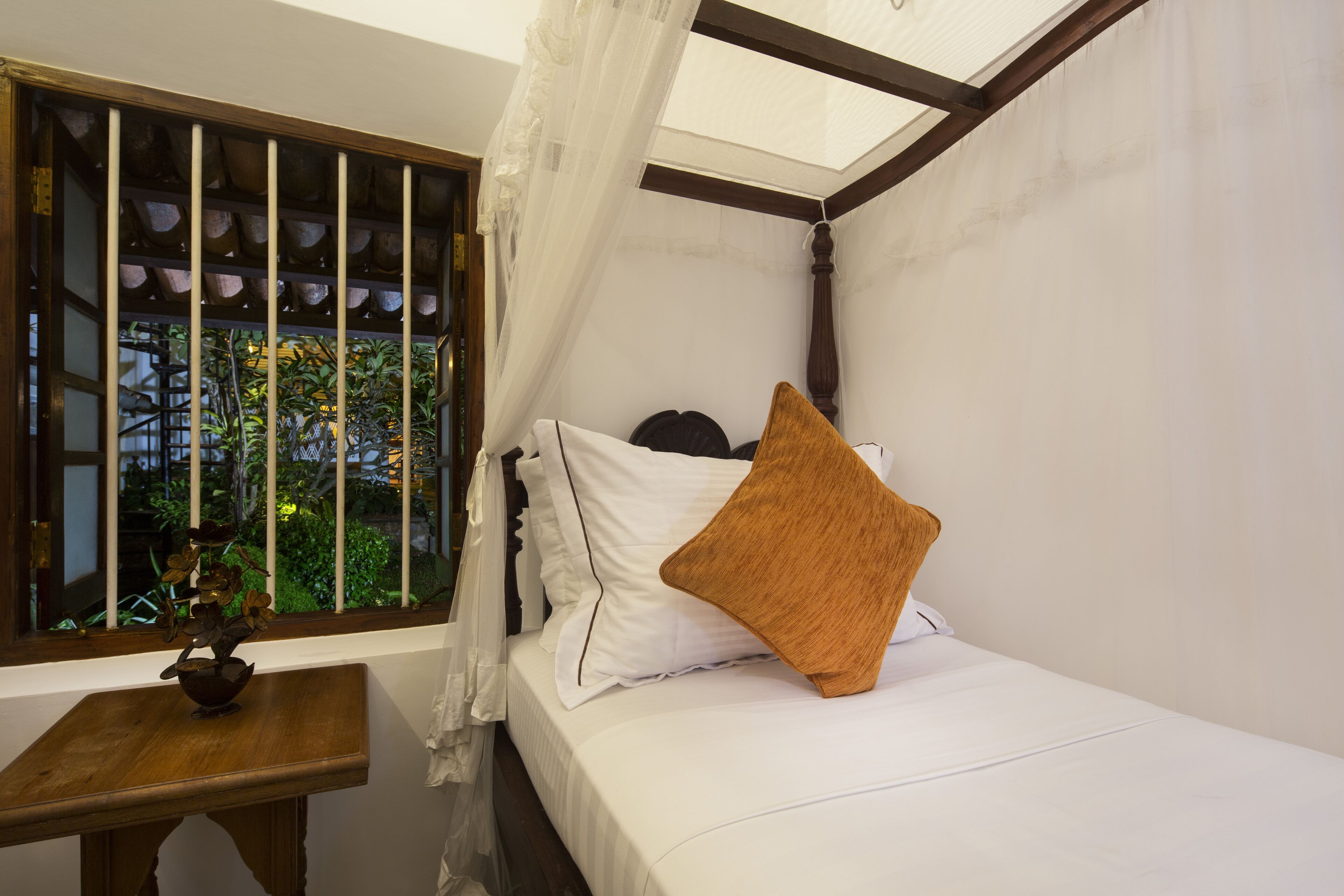 A single bed with white bedding and a brown pillow, a small wooden table with a vase, and a window with barred glass and a view of greenery.