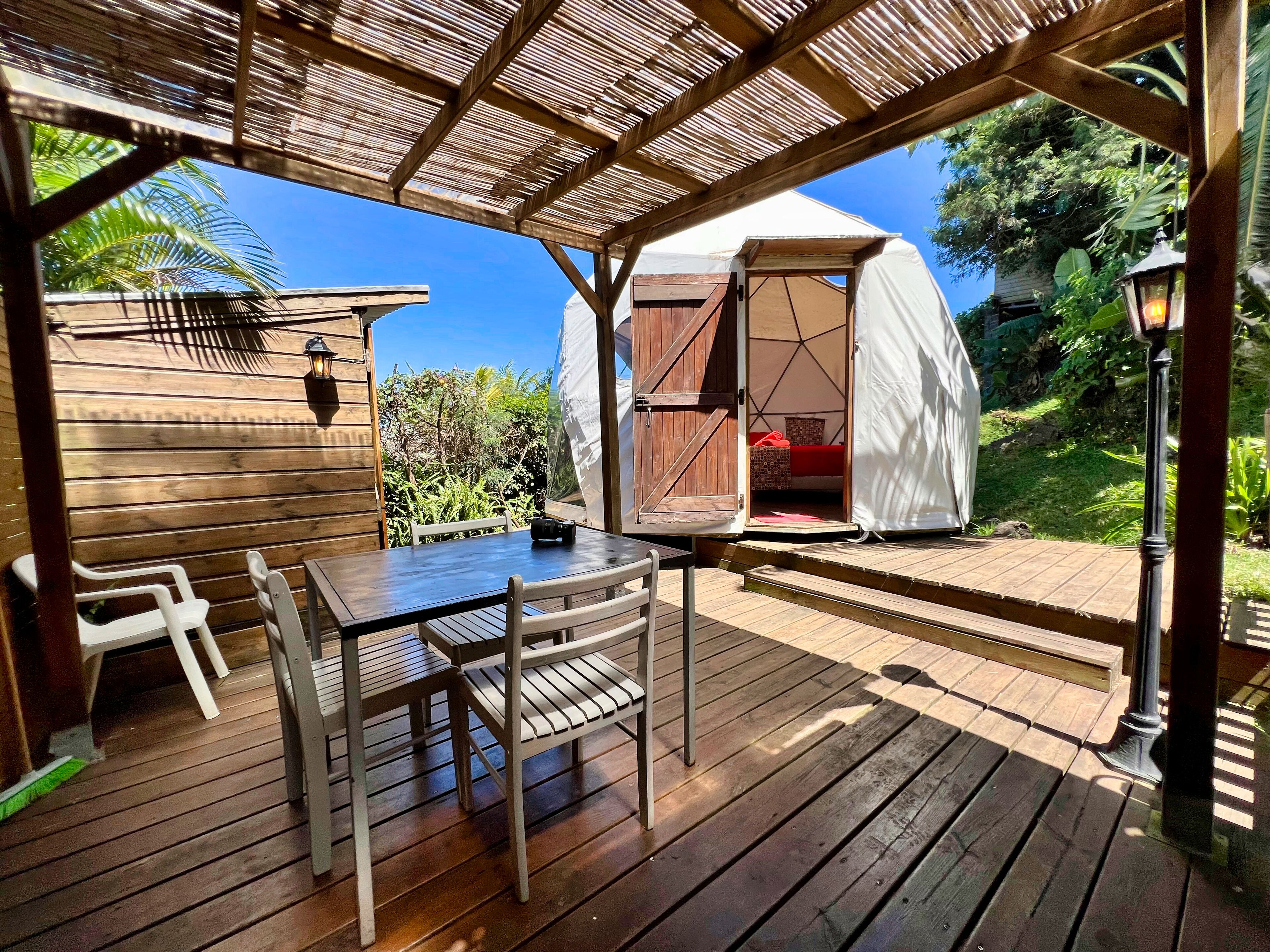 Una terraza de madera con mesa y sillas, un espacio cubierto con una carpa y vistas a la vegetación.