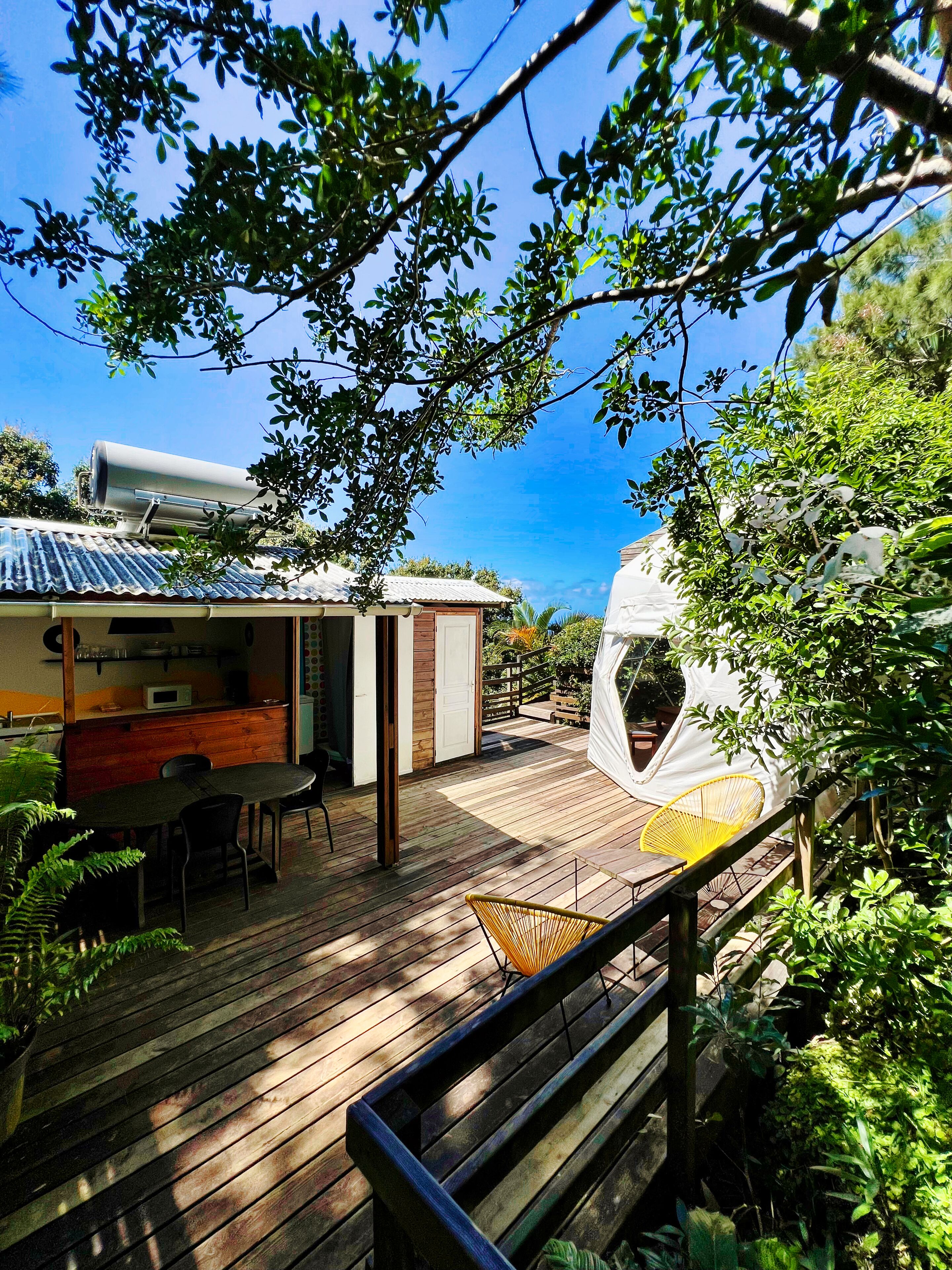 Une terrasse en bois avec un espace repas, une tente blanche et un réservoir d’eau en métal.