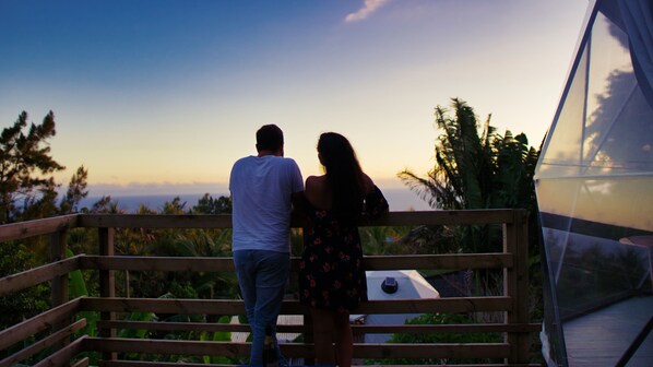 A couple standing on a wooden deck overlooking a scenic view with trees and a clear sky.