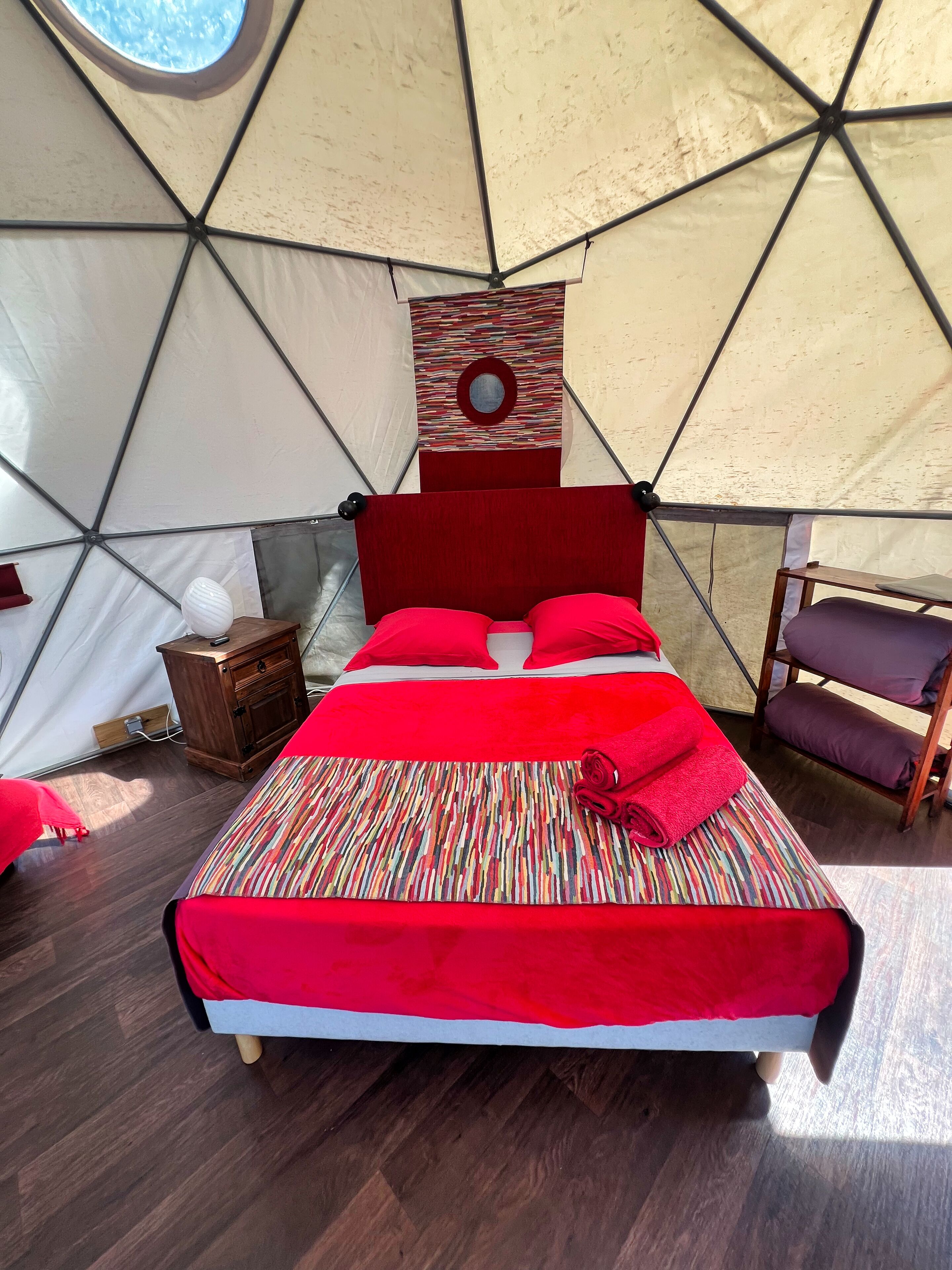 Una cama con ropa de cama roja en un espacio tipo tienda, con una mesita de noche de madera y una pequeña mesa.