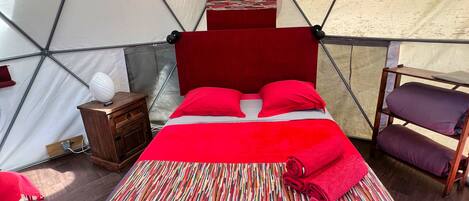 Una cama con ropa de cama roja en un espacio tipo tienda, con una mesita de noche de madera y una pequeña mesa.