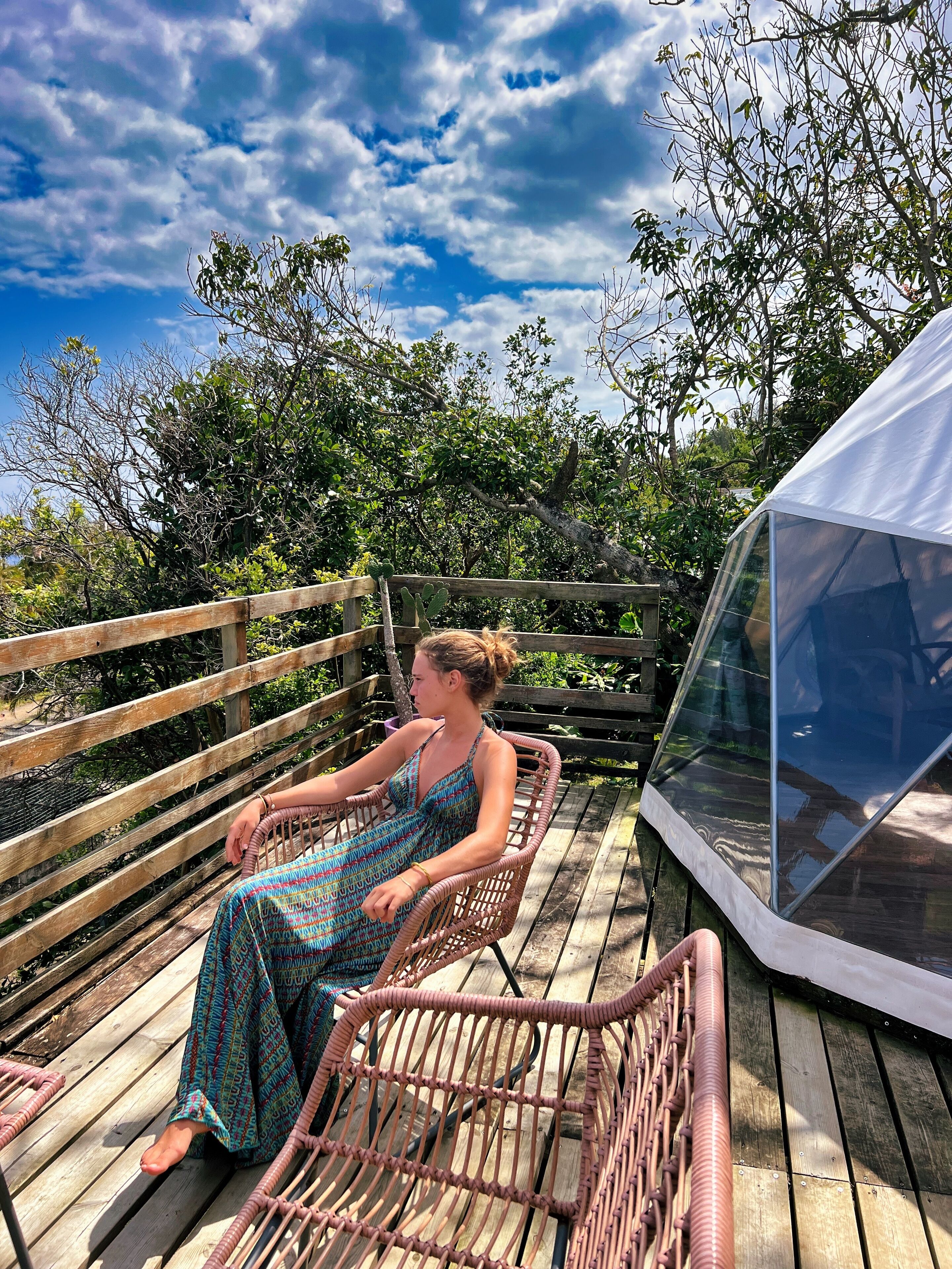 Une personne est assise sur une chaise en osier, sur une terrasse en bois, et une tente transparente se trouve à proximité.