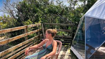 Une personne est assise sur une chaise en osier, sur une terrasse en bois, et une tente transparente se trouve à proximité.