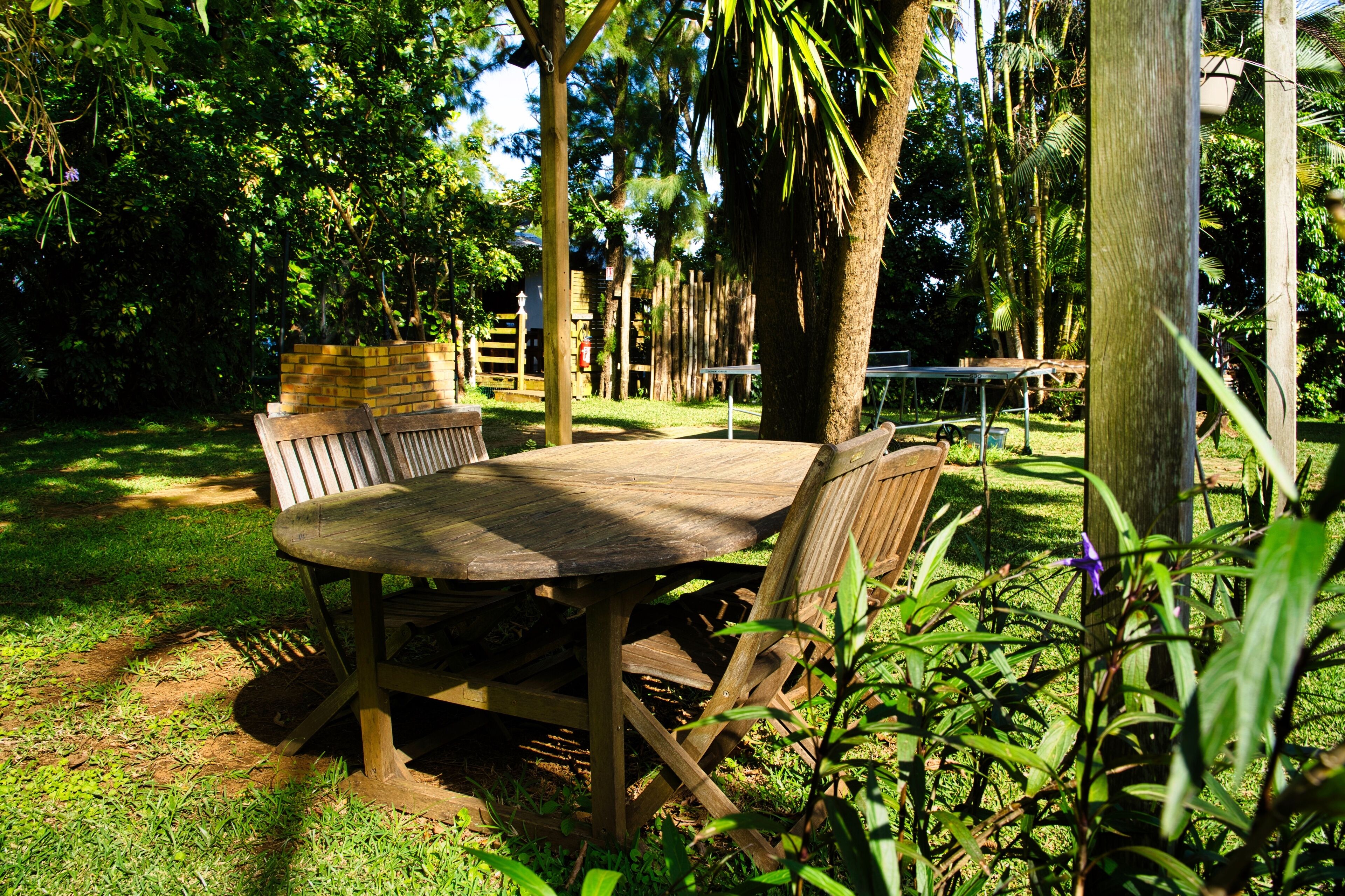 Una mesa de madera y bancos en un jardín con árboles y un muro de ladrillo al fondo.