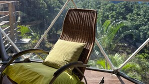 A wicker chair with a green cushion on a balcony with a view of a lush green landscape and a clear blue sky. - Bubble Dome Village- Glamping (Saint-Joseph)