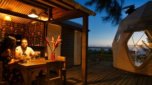 A wooden deck with a dining table and chairs, a tent with a circular window, and a view of the ocean. - Bubble Dome Village- Glamping (Saint-Joseph)