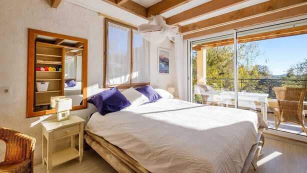 A bedroom with a large bed, a wooden nightstand, a wicker chair, and a balcony with a table and chairs. - Large Provencal house with magnificent views in the heart of a historic village (Fayence)