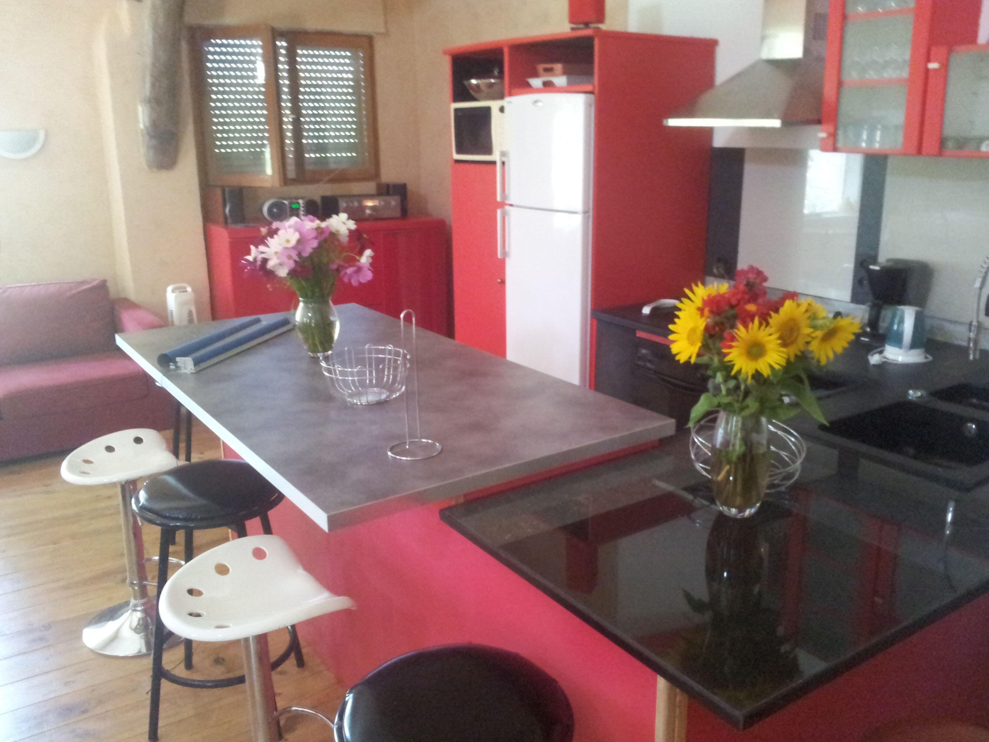 Cuina amb un frigorífic vermell i blanc, una cafetera i un ram de girassols. Hi ha una taula de dinar amb dues cadires blanques i un altre ram amb flors roses.