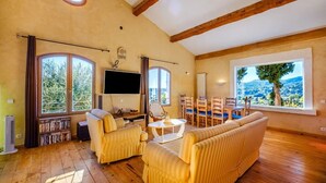 A spacious living room with wooden floors, large windows offering a view of the countryside, a flat-screen TV, and a dining area with wooden chairs. - Large Provencal house with magnificent views in the heart of a historic village (Fayence)