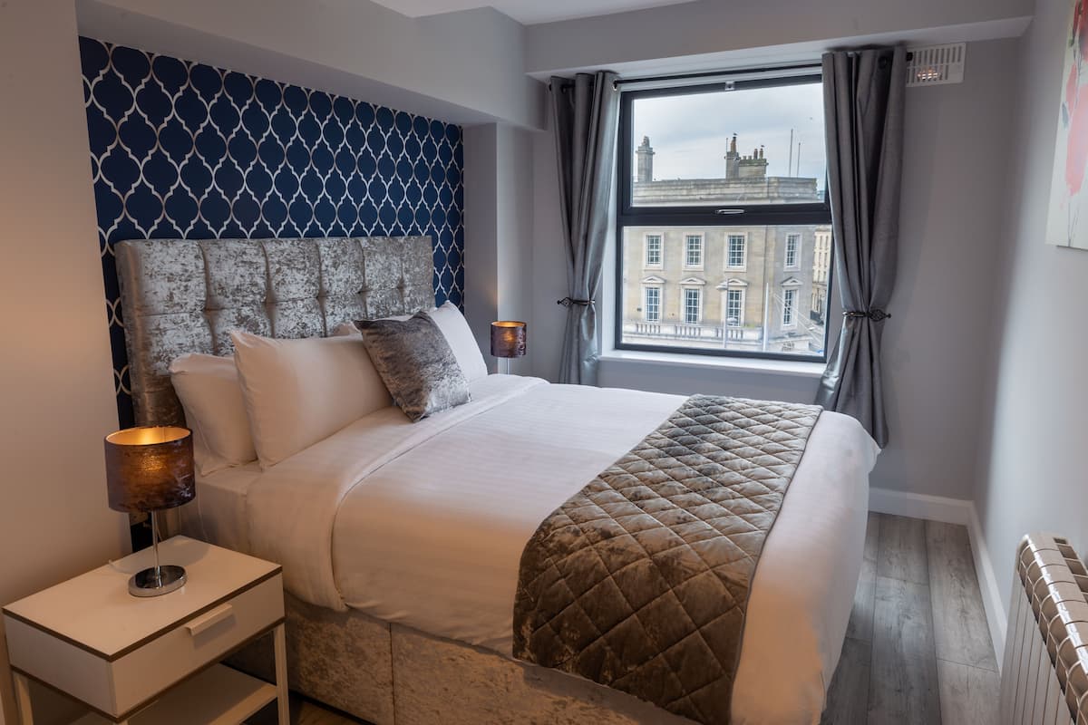 A neatly made bed with a quilted blanket, a bedside lamp, and a view of a building through a window with curtains.