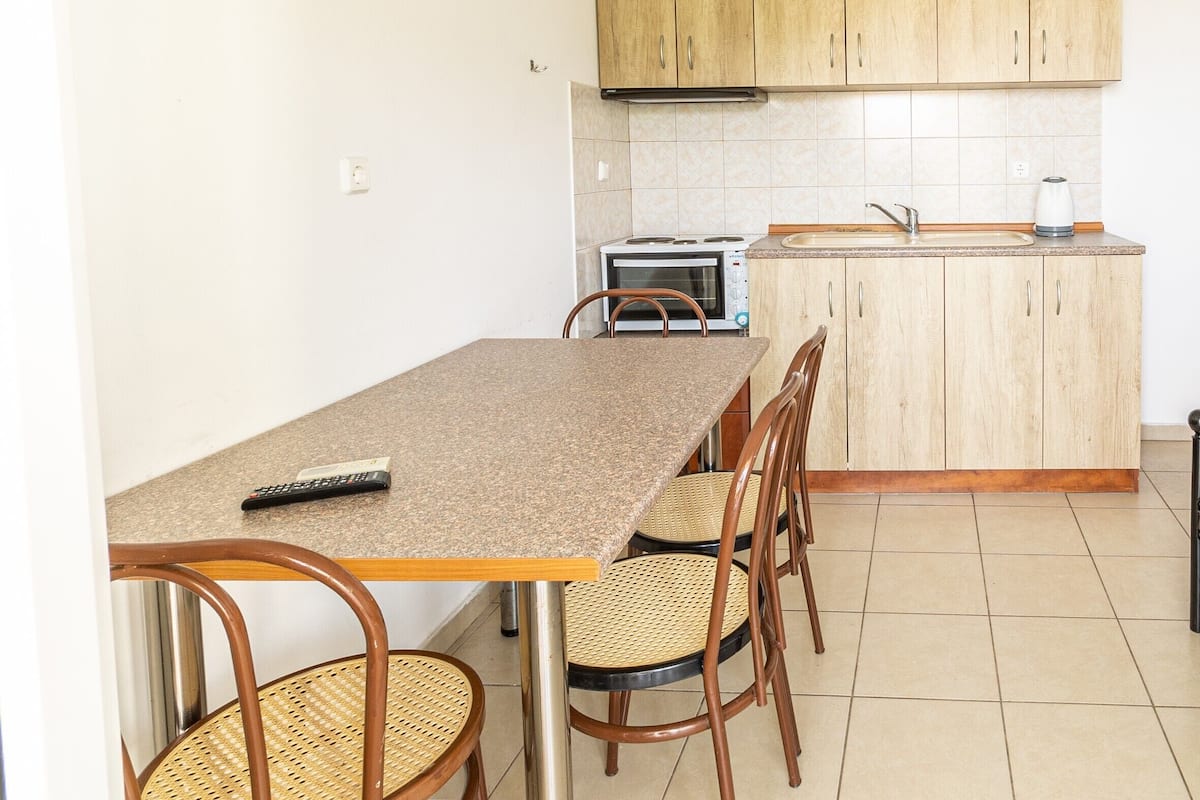 A small kitchen with wooden cabinets, a microwave, a kettle, and a dining table with chairs.