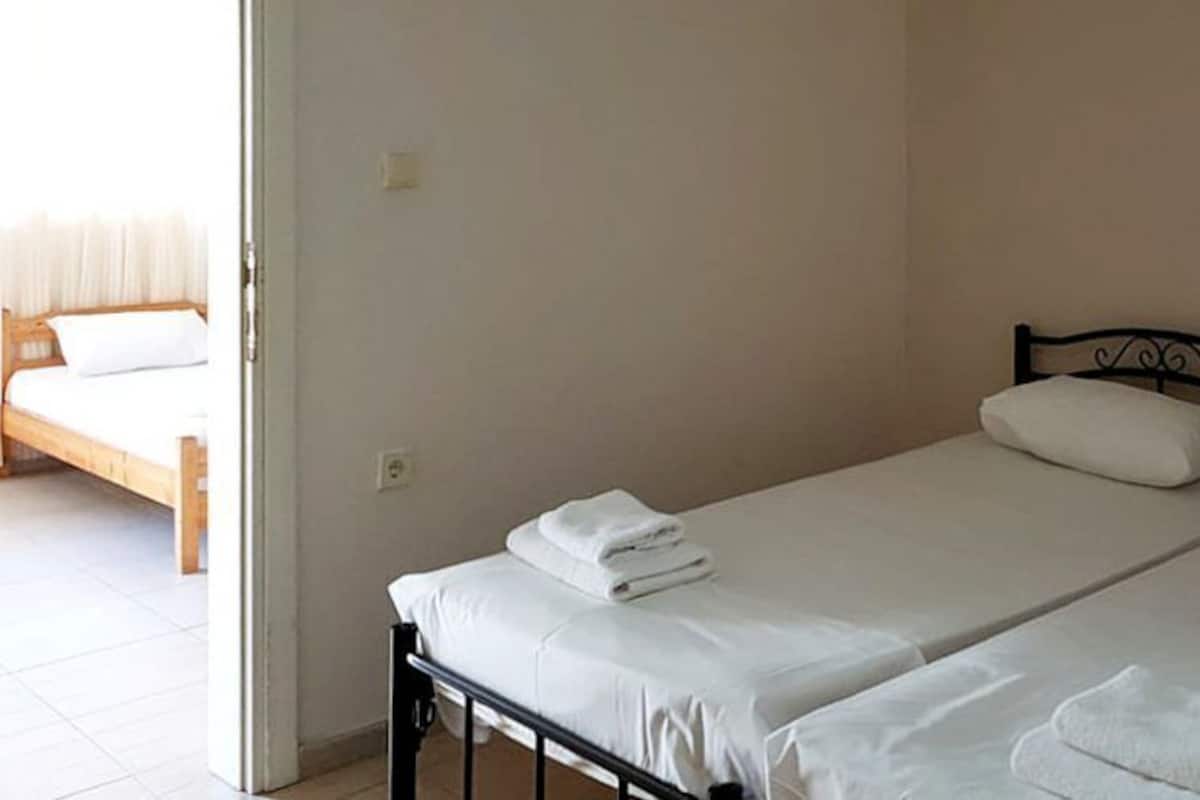 A room with two single beds, each with white linens and pillows, a wooden bed frame, and a plain wall.