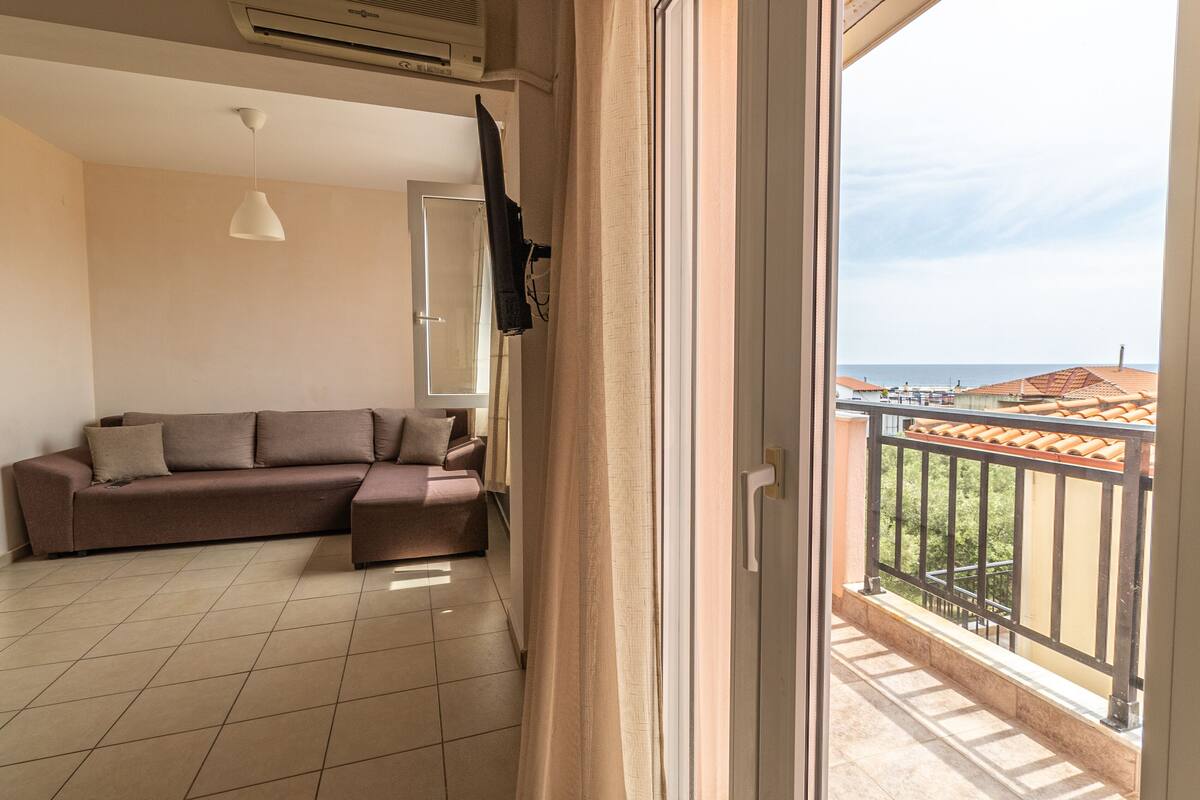 A living room with a sofa, a hanging light fixture, and a view of the sea through a balcony.