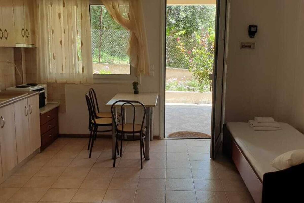 A compact kitchen with a dining table and chairs, a small window with curtains, and a single bed with a mattress and pillows.