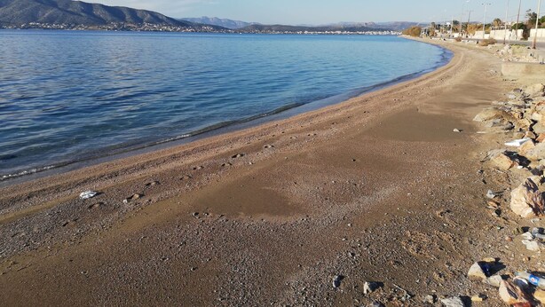 A pebbly beach with a clear blue sea, a distant mountain range, and a row of street lamps along the shore. - Salamina's Apartment (Salamis)