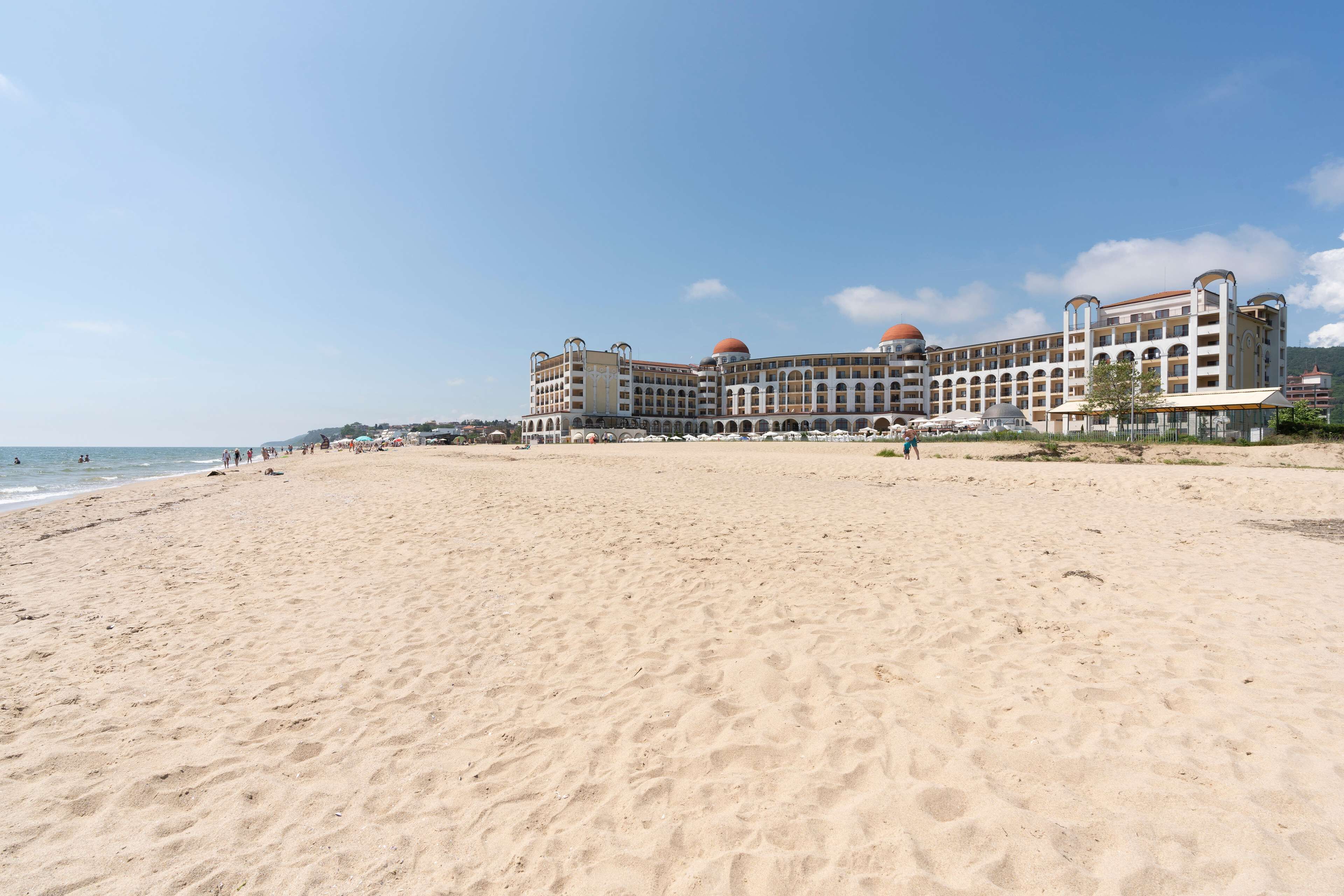 A large hotel with multiple floors and domed roofs, situated on a sandy beach with the sea in the background.