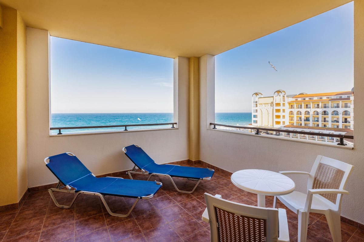 A balcony with two blue lounge chairs, a small round table, and a white chair. The balcony overlooks a clear blue sea and a building with multiple balconies.