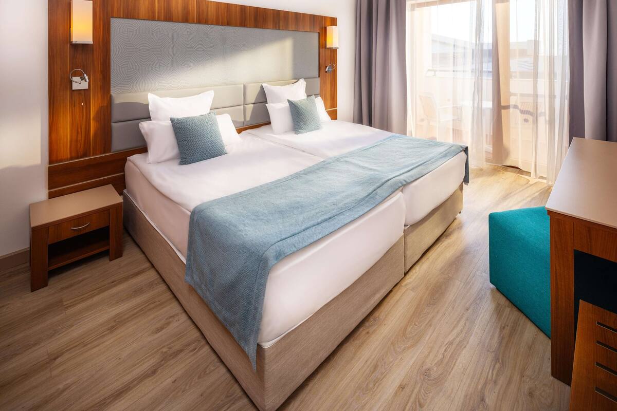 A hotel room with a large bed, wooden headboard, bedside tables, and a view of the outdoors through large windows.