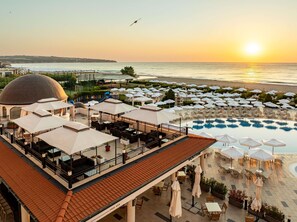 A rooftop pool area with lounge chairs and umbrellas, overlooking the sea at sunset.