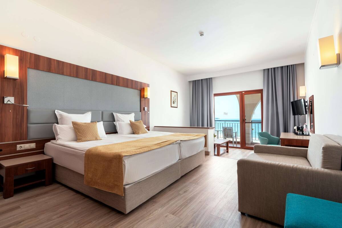 A hotel room with a large bed, a sofa, a desk, and a balcony with curtains.