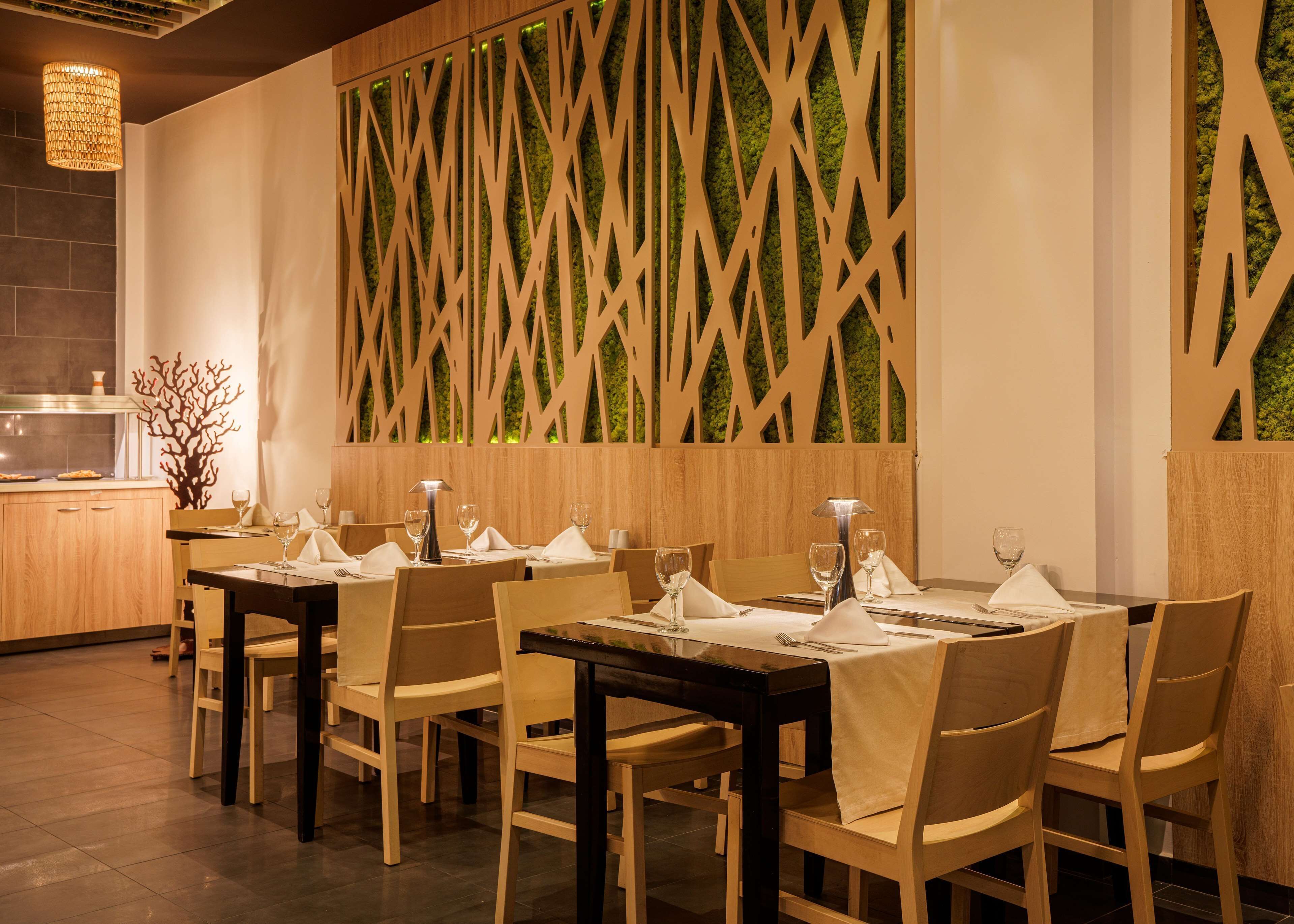 Interior de restaurante con mesas y sillas de madera, pared decorativa con motivo geométrico y una pequeña cocina visible a la izquierda.