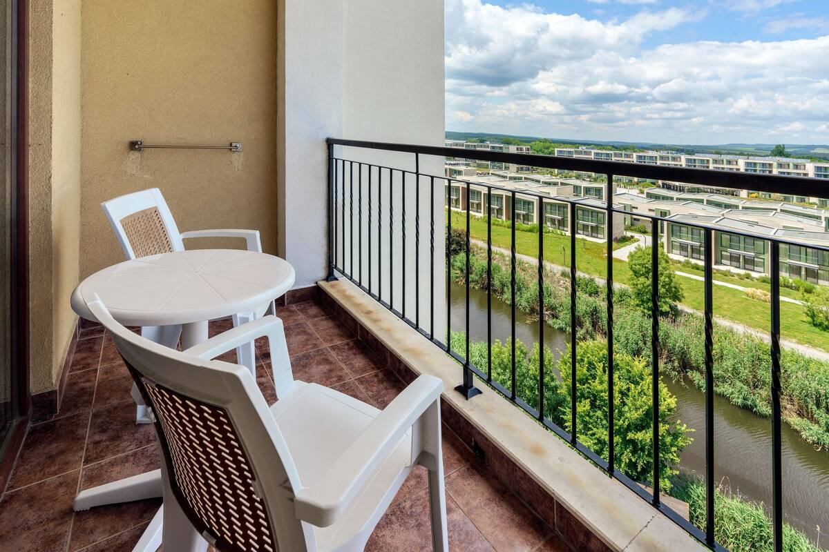 A balcony with a table and chairs, a view of a river, and modern buildings in the background.