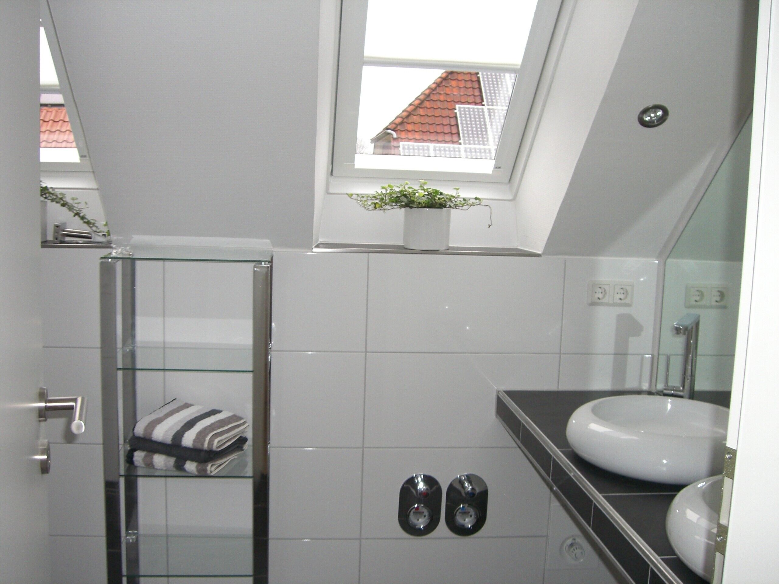 A modern bathroom with a glass shower enclosure, a white sink, and a small window with a view of a red-roofed building.