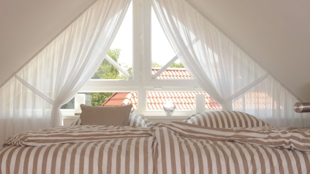 Ein Schlafzimmer mit schrägem Dach, einem Bett mit gestreifter Bettwäsche und transparenten Vorhängen.