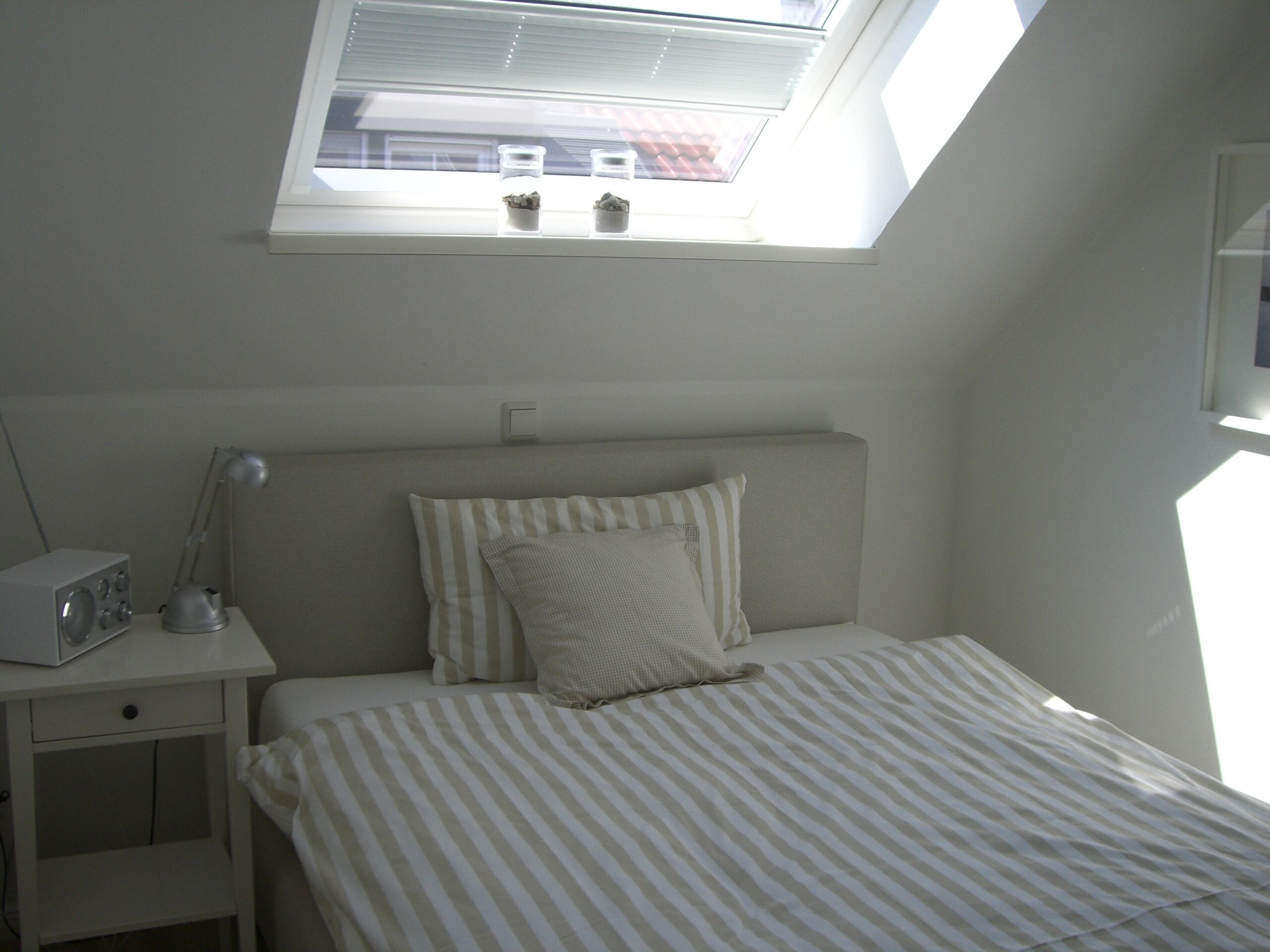Un dormitorio con tragaluz, una cama con ropa de cama a rayas, una mesita de noche con lámpara y una radio pequeña.