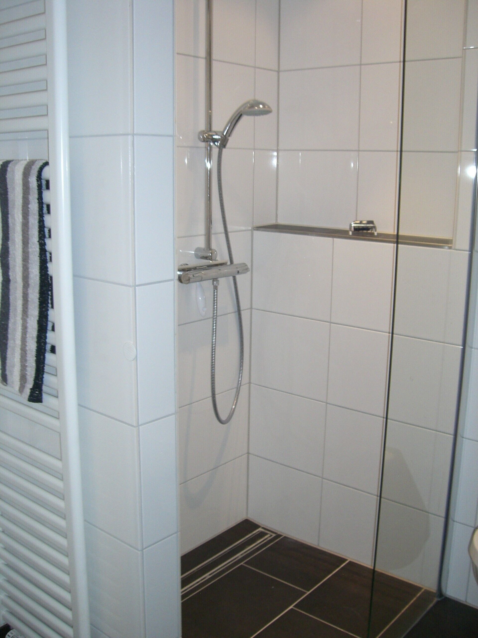 A bathroom with white tiled walls, a shower with a chrome faucet and hose, a towel rack with a striped towel, and a dark brown tiled floor.