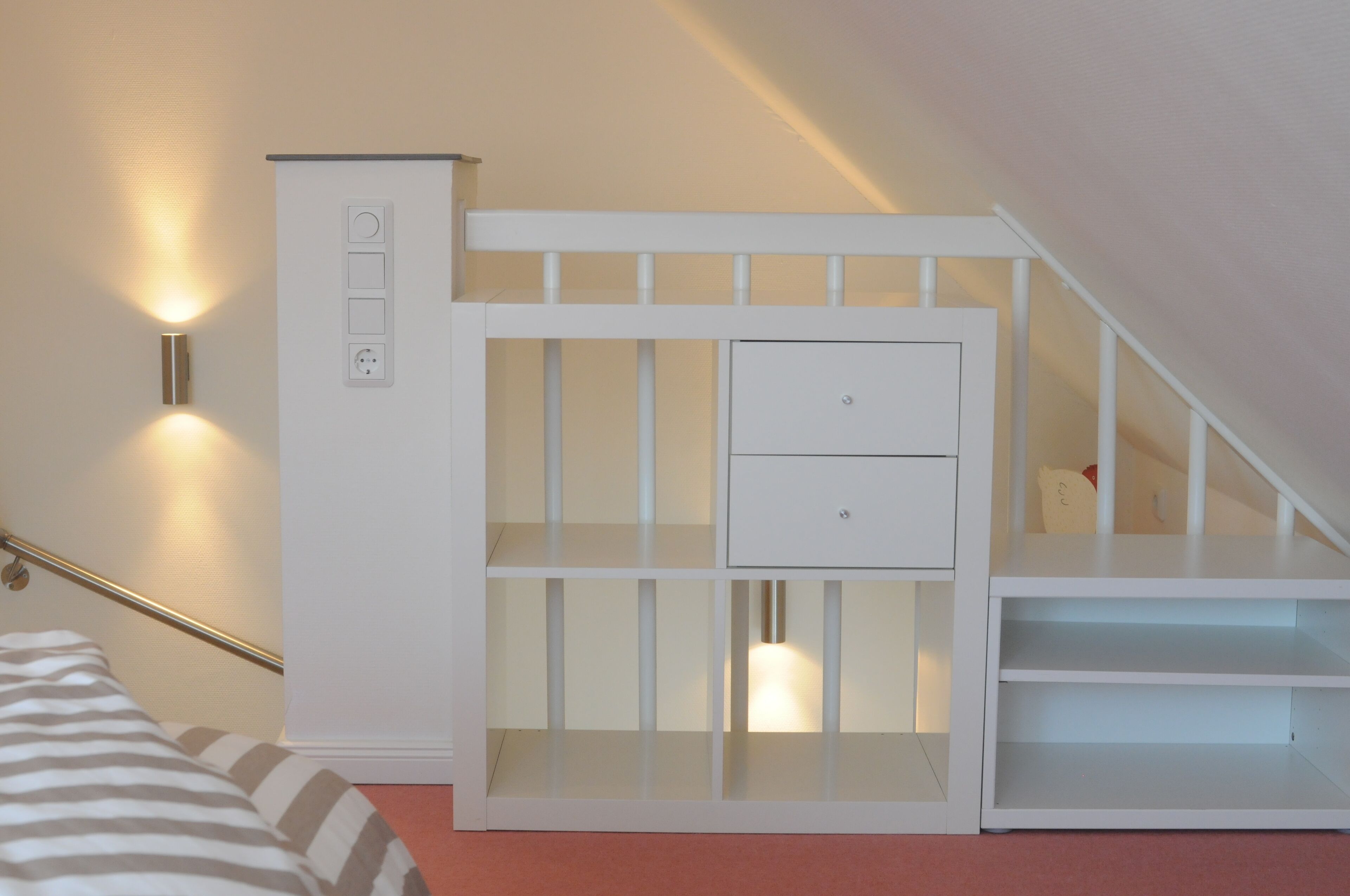 Una habitación blanca de varios niveles, con una escalera, una cama con ropa de cama a rayas y un aplique de pared.