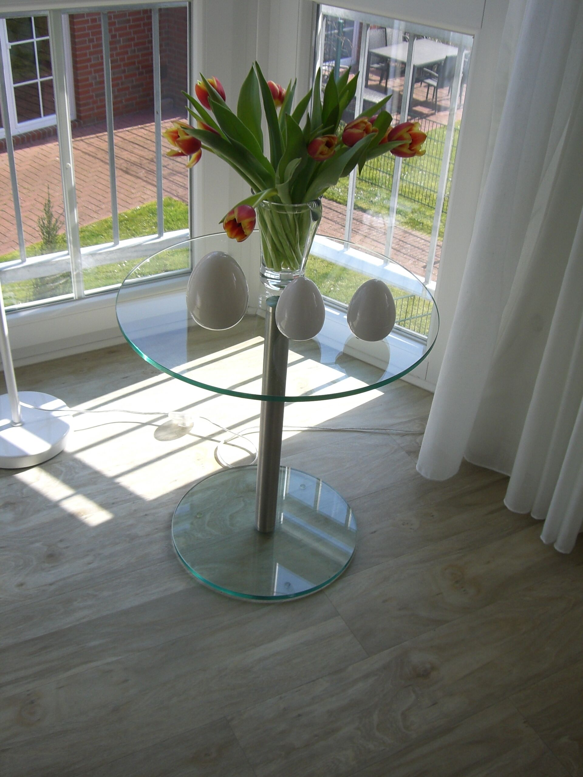 A glass table with a vase of tulips and three spherical objects on a wooden floor near a window.