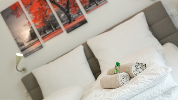 A neatly made bed with white linens and rolled towels, a green bottle, and a framed picture of red trees on the wall. - Apartment Duomo (Bolzano)