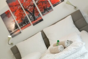 A neatly made bed with white linens and rolled towels, a green bottle, and a framed picture of red trees on the wall. - Apartment Duomo (Bolzano)