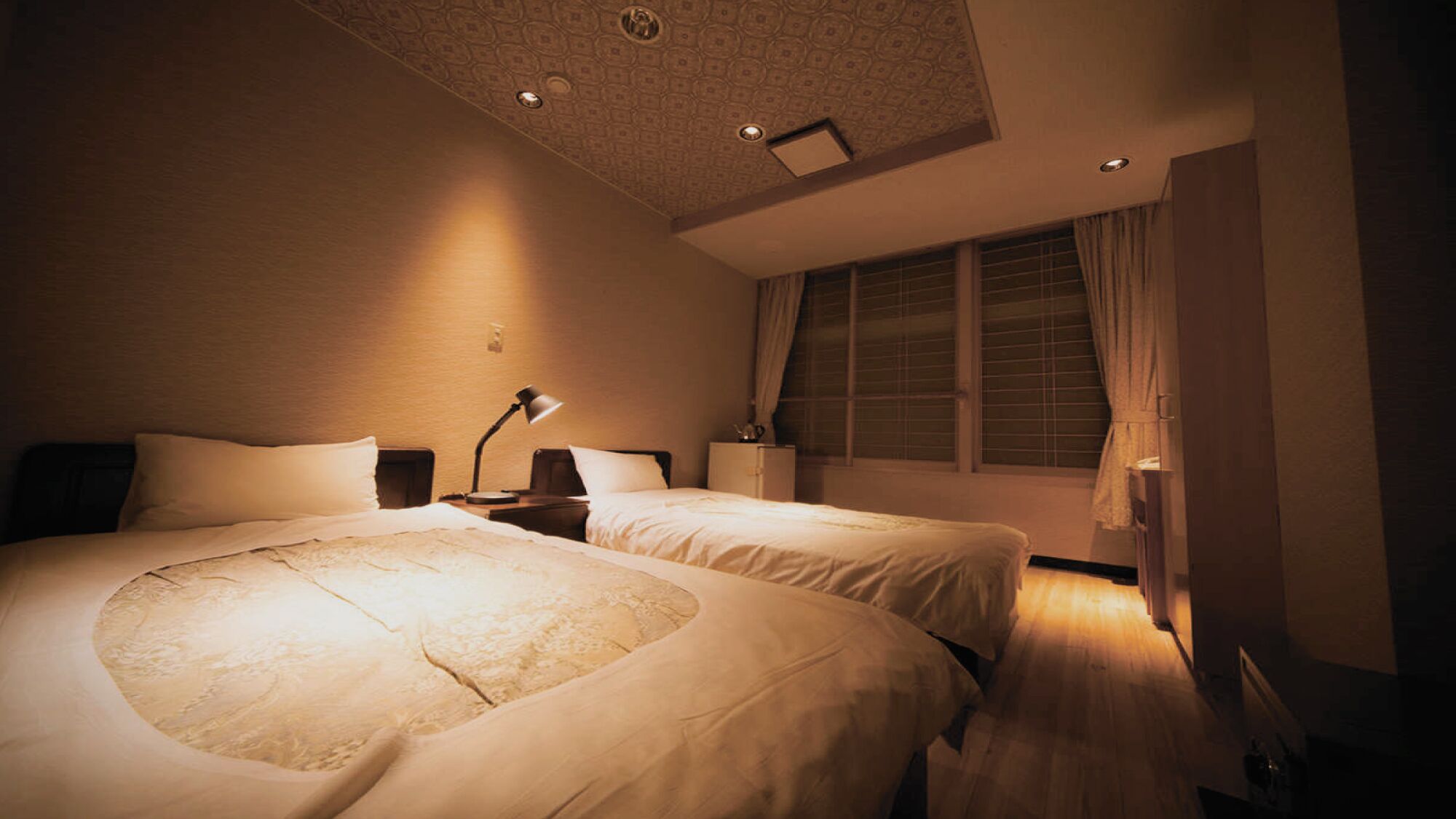 Ein Hotelzimmer mit zwei Betten, einem Schreibtisch, einer Lampe, einem Fenster mit Vorhängen und einer gemusterten Decke.