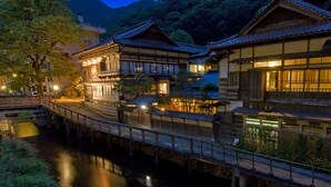 Un ensemble de bâtiments traditionnels japonais comprenant plusieurs structures reliées par un pont qui enjambe un cours d’eau.