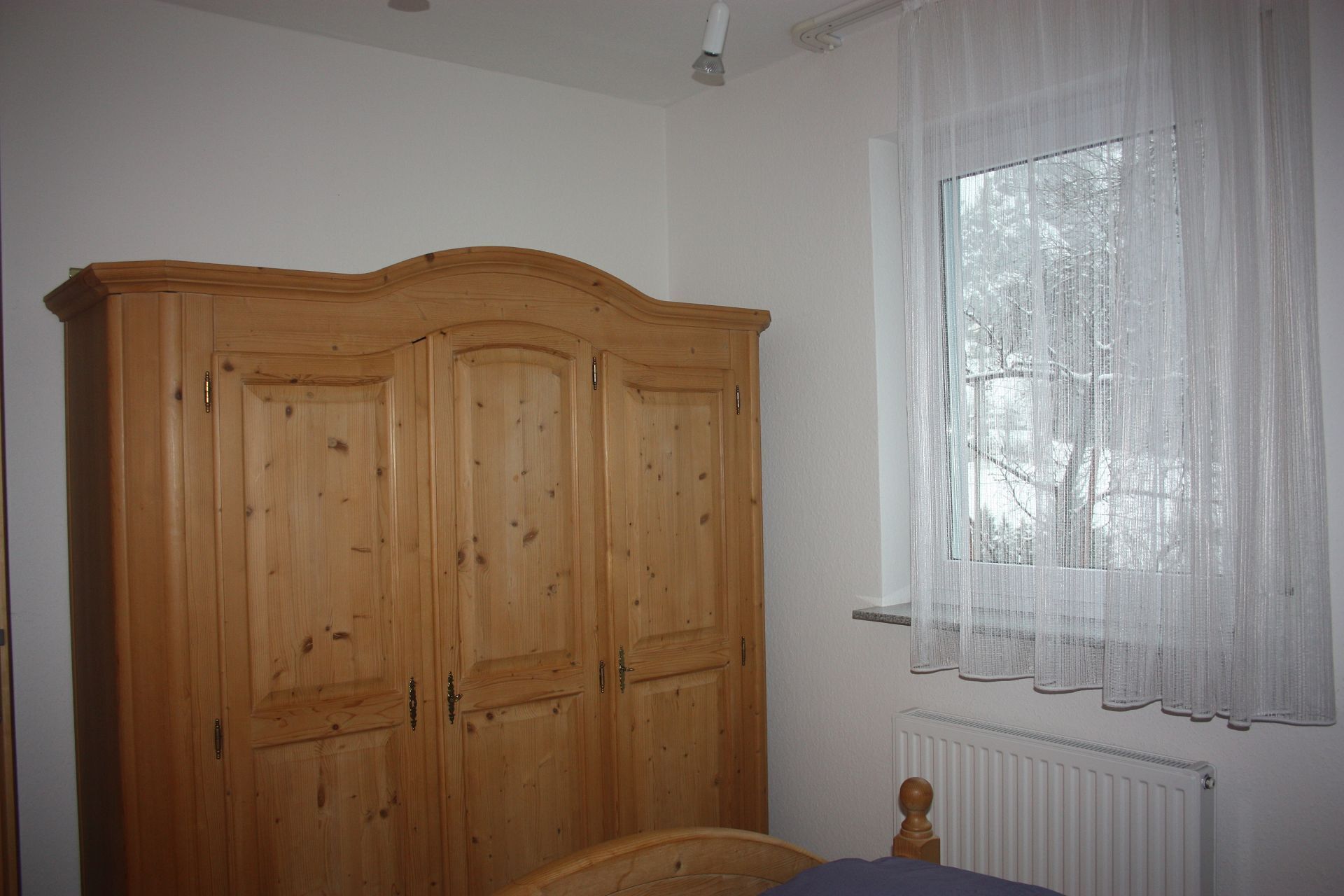 A bedroom with a wooden wardrobe, a window with sheer curtains, and a radiator.