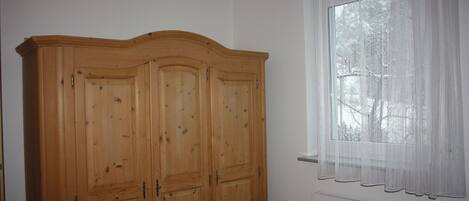 A bedroom with a wooden wardrobe, a window with sheer curtains, and a radiator.