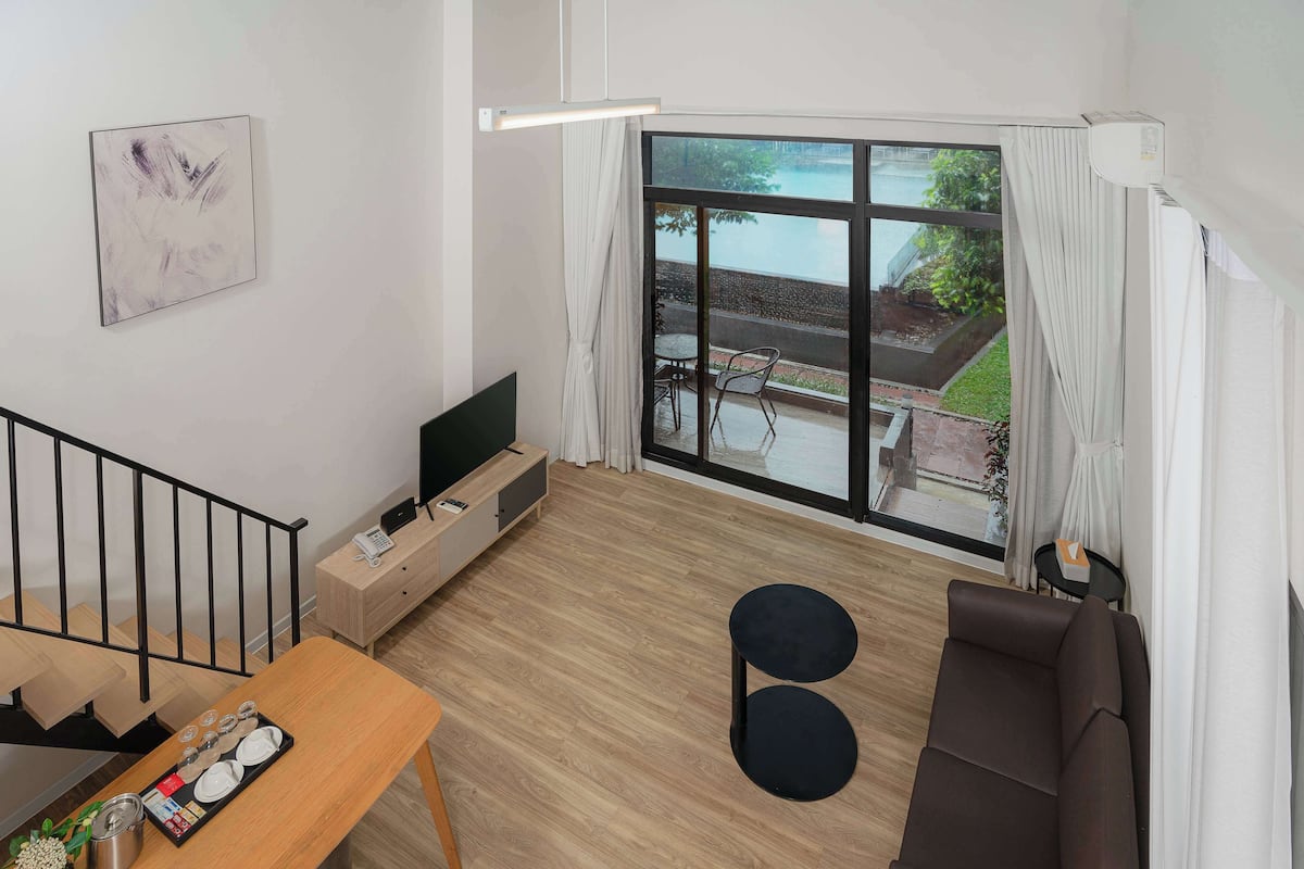 A modern living room with a wooden floor, a brown sofa, a dining table with a tray of dishes, a TV on a wooden stand, and a large window with a view of a garden and pool.