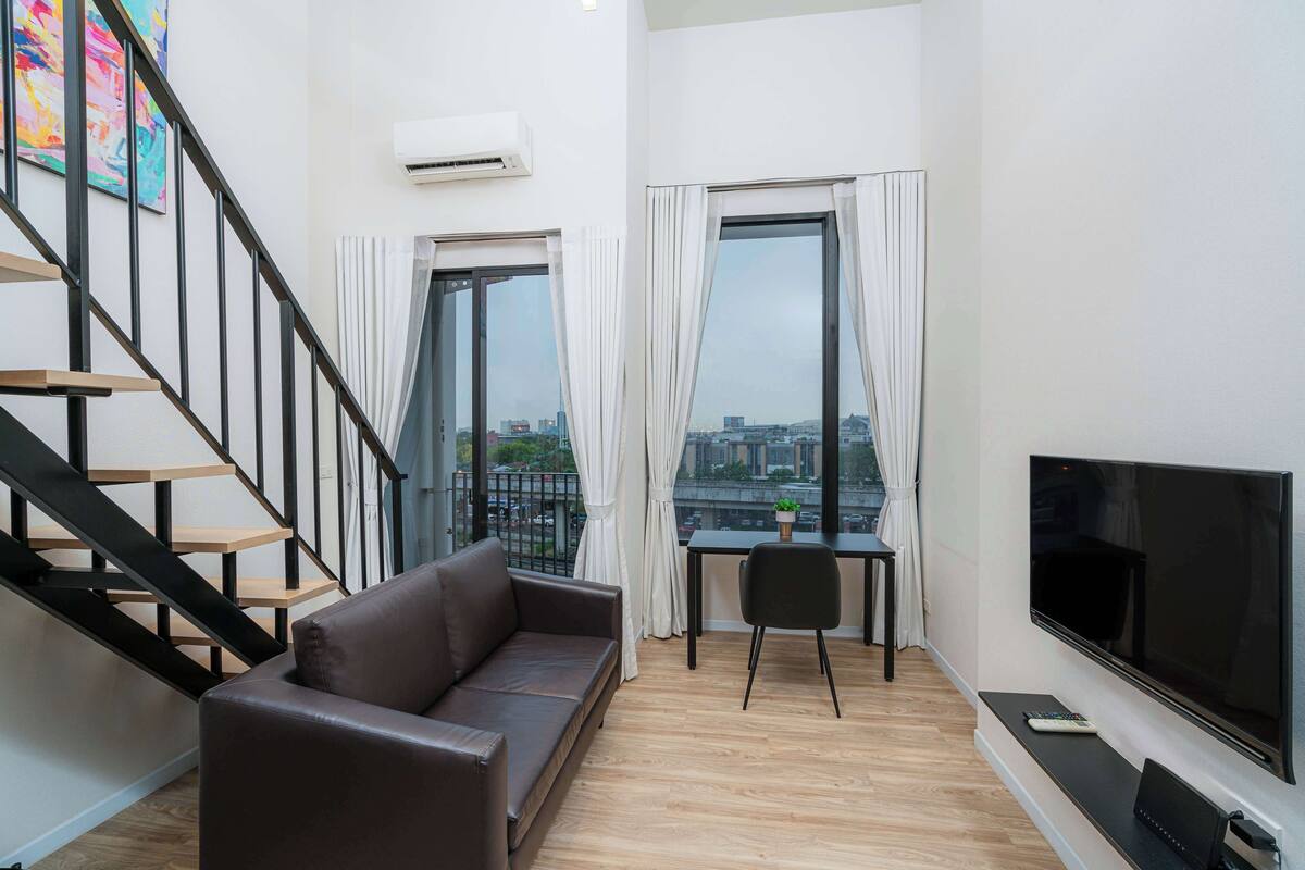 A modern living room with a brown leather sofa, a flat-screen TV, a dining table with a chair, and a staircase with black railings.