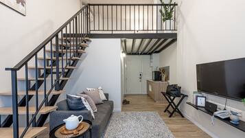 Una sala de estar moderna con una escalera, un sofá gris, una mesa de centro, un televisor y una pequeña cocina.