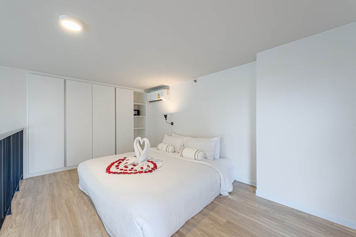 A modern bedroom with a large bed, white bedding, a heart-shaped decoration on the bed, a built-in wardrobe, and a wall-mounted air conditioning unit.
