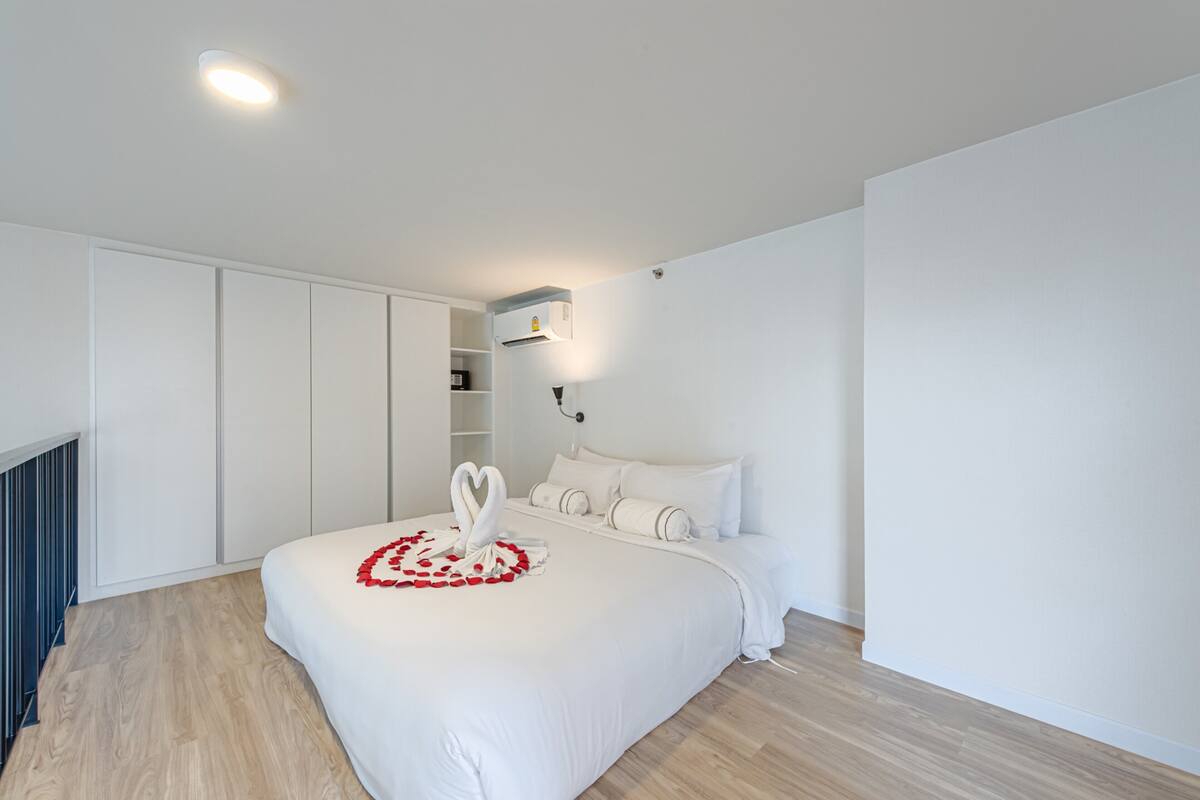Un dormitorio moderno con una cama grande, ropa de cama blanca, una decoración con forma de corazón sobre la cama, un armario empotrado y un aire acondicionado instalado en la pared.