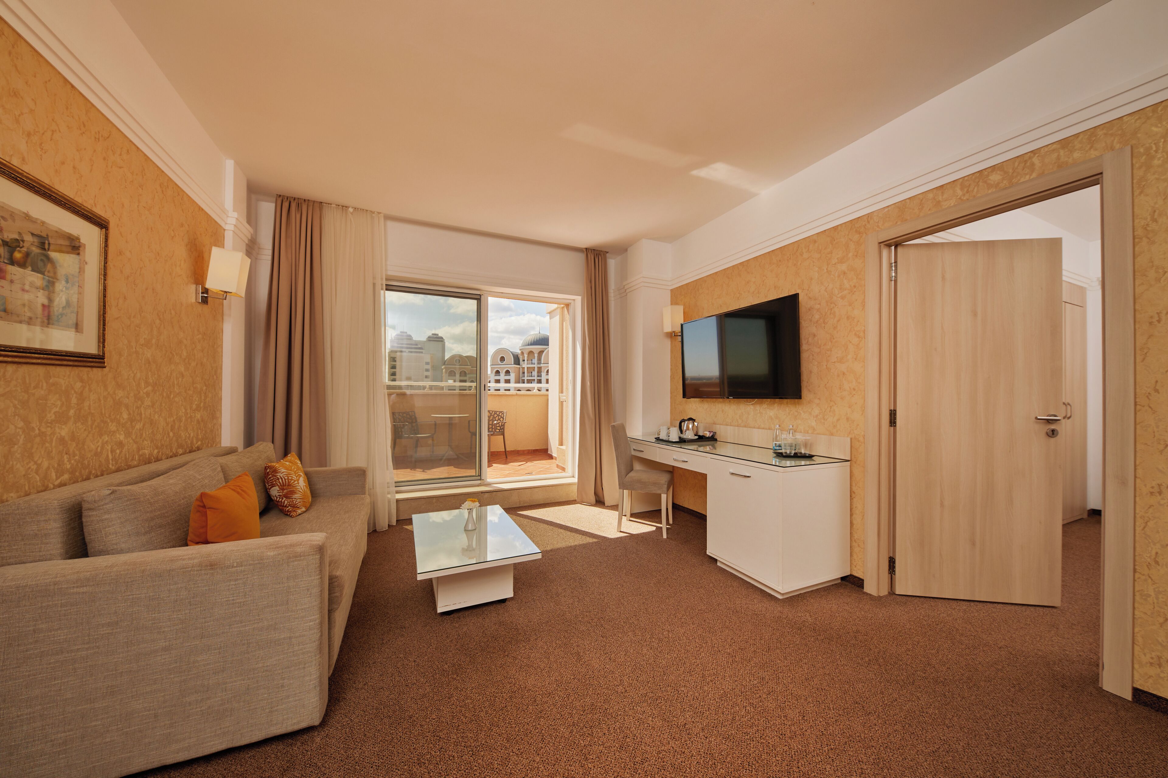 Ein Hotelzimmer mit einer Sitzecke, einem kleinen Couchtisch, einem Schreibtisch mit Stuhl, einem Flachbildfernseher und einer Schiebetür zu einem Balkon mit Blick auf die Stadt.