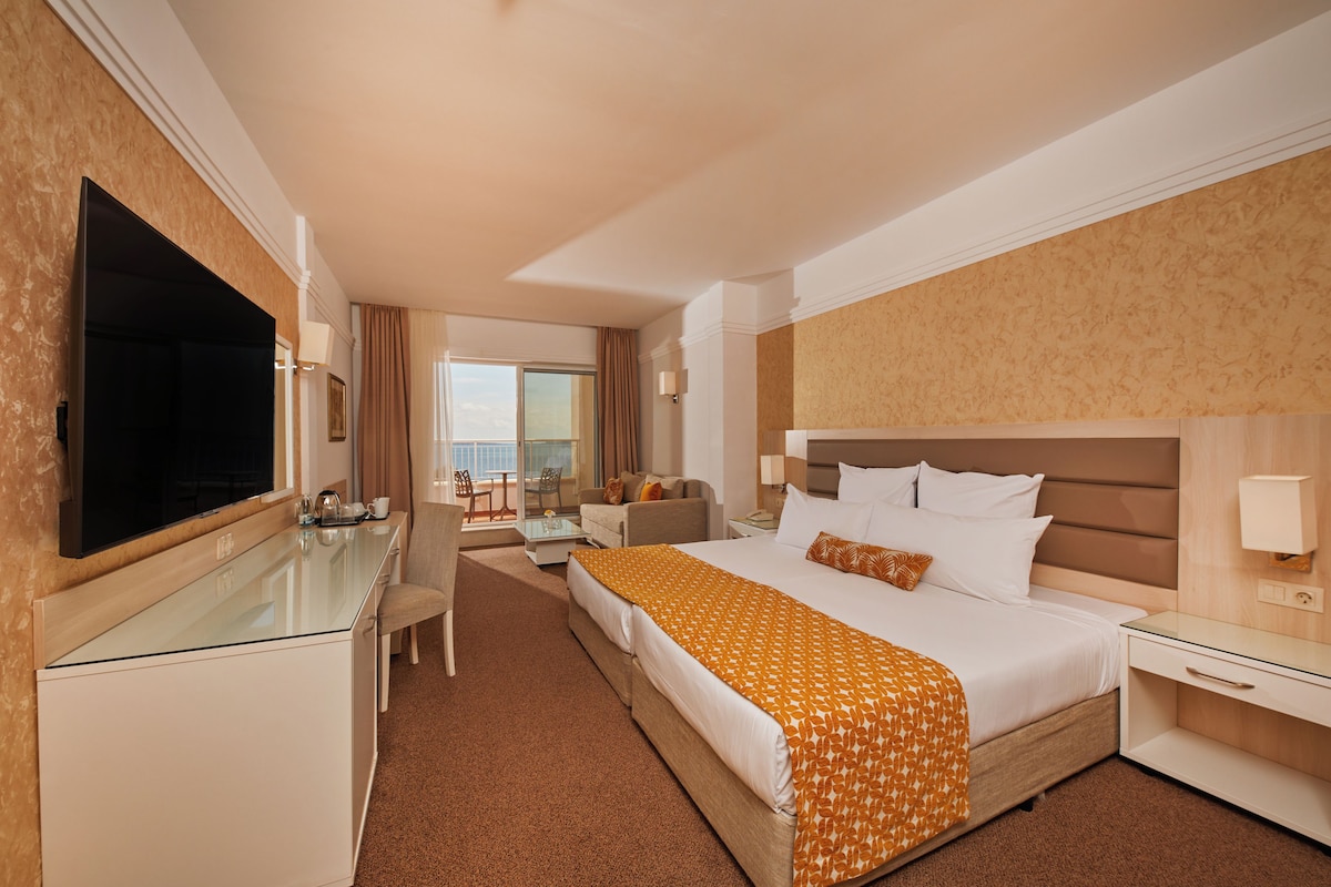 Une chambre d’hôtel dotée d’un grand lit, d’un bureau avec un téléviseur à écran plat, d’un coin salon avec un canapé et des fauteuils, et offrant une vue sur la ville par la fenêtre.