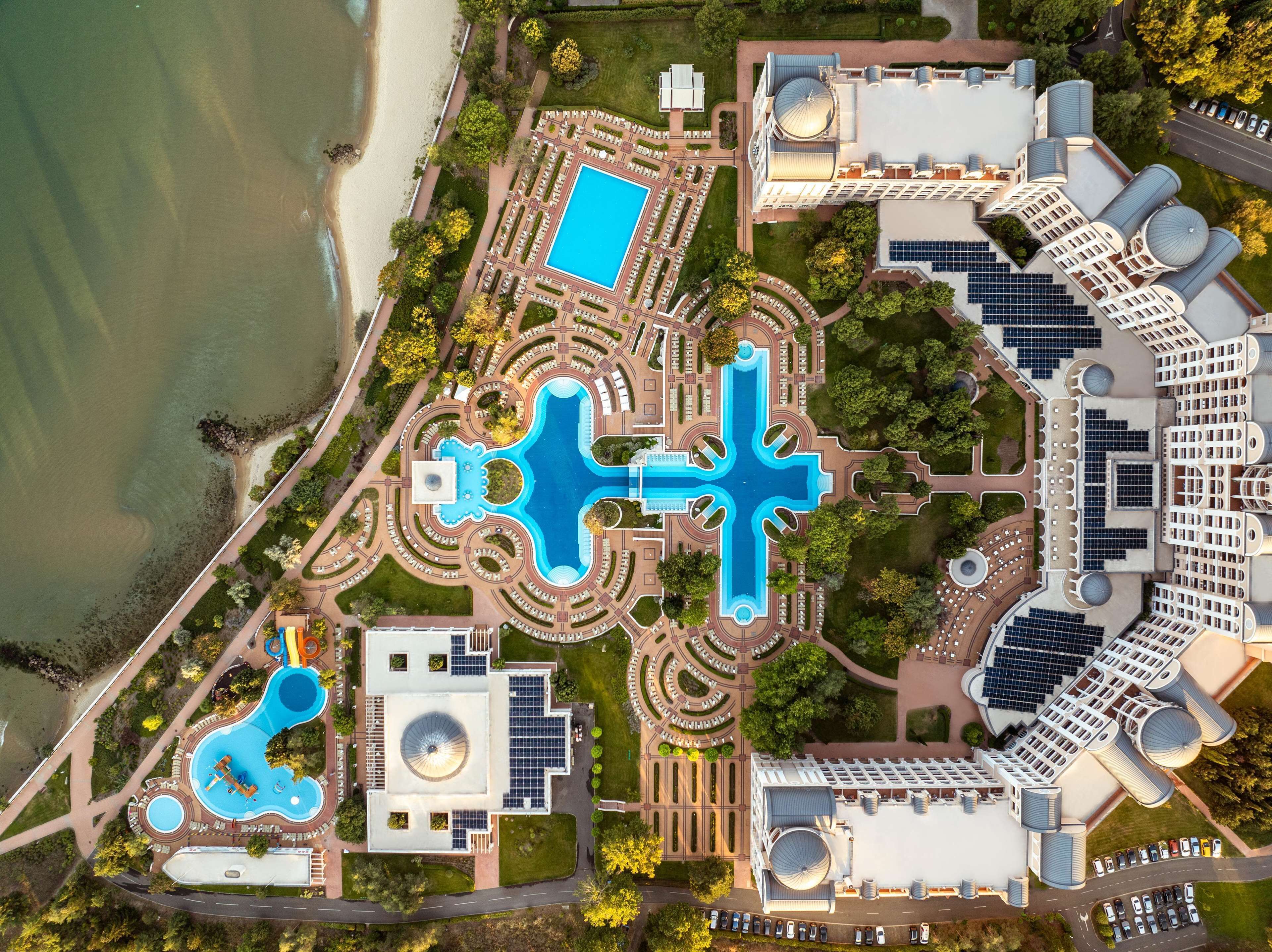 Vista aérea de un resort con múltiples piscinas, una piscina central grande y un estacionamiento con paneles solares.
